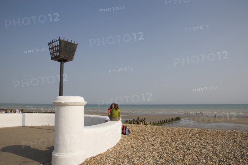 ENGLAND, West Sussex, Bognor Regis, Two young women sitting on a white wall facing each other talking near a torch beacon. Sand and shingle beach with families seen at the waters edge behind