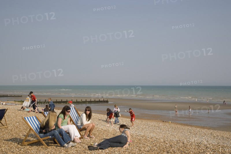 ENGLAND, West Sussex, Bognor Regis, A group of girls sitting on deck chairs on shingle beach with children playing behind