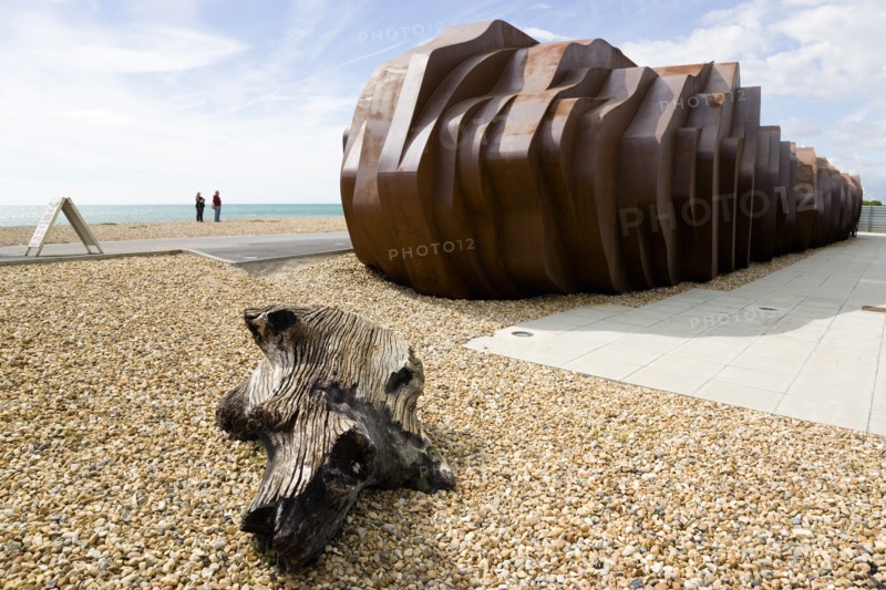 ENGLAND, West Sussex, Littlehampton, Two people standing on the beach looking at the rusted metal structure of the fish and seafood restaurant the East Beach Cafe designed by Thomas Heatherwick with an A-frame on the promenade and driftwood on the pebble beach in the foreground