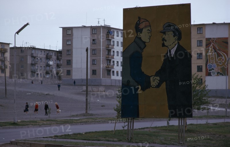 MONGOLIA, Ulan Bator, Soviet billboard in central Ulan Bator  depicting Lenin shaking hands with Mongolia's first president Sukhebator in urban area with multi-storey apartment blocks behind and people crossing a central square . Ulaanbaatar East Asia Soviet Mongolia Asian Baator Male Men Guy Mongol Uls Ulaan