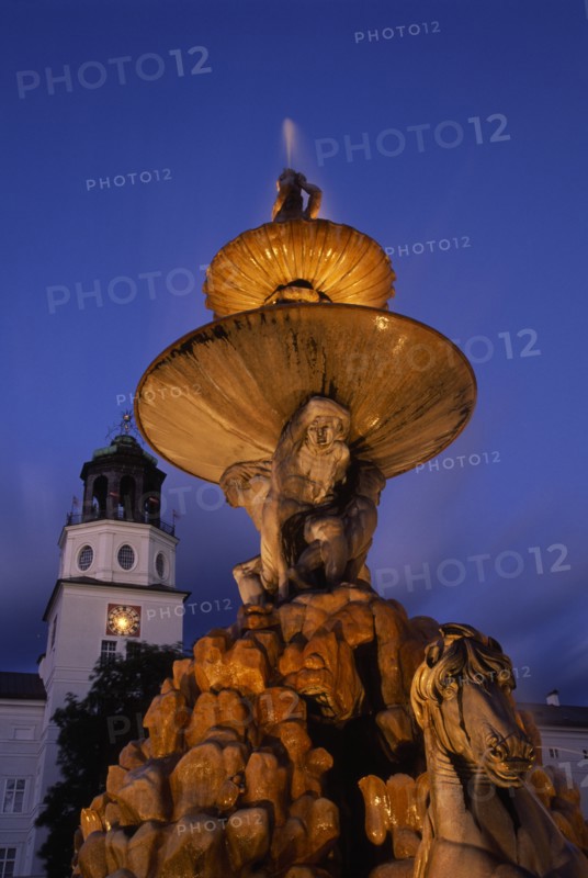 AUSTRIA, Salzburg, Part view of Baroque fountain with statues of horse and contorted figures and the Glockenspiel Carillon tower behind at night.