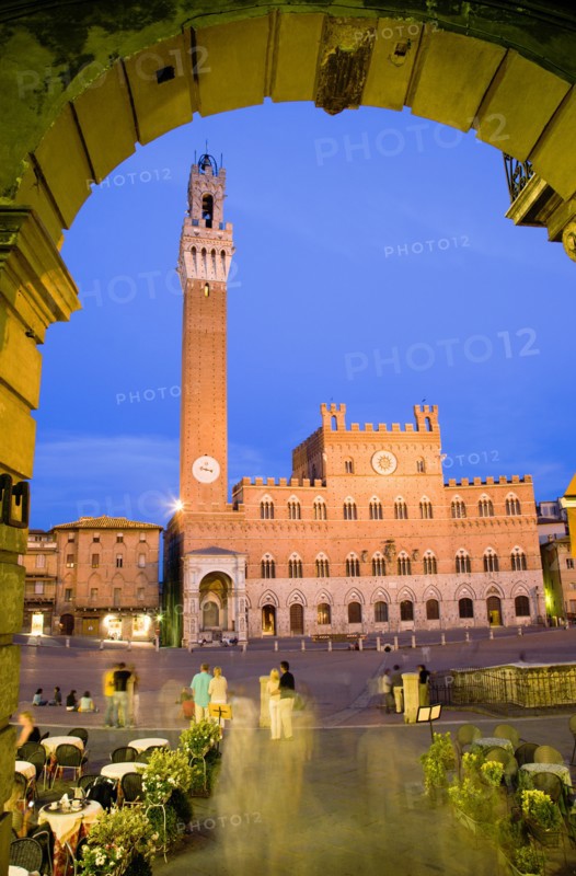 ITALY, Tuscany, Siena, "Early evening in the Piazza del Campo seen through an archway with people at restaurant tables and walking in the square. The illuminated Torre del Mangia, the second tallest belltower in Italy, is beside the Palazzo Publico at the far side of the piazza"