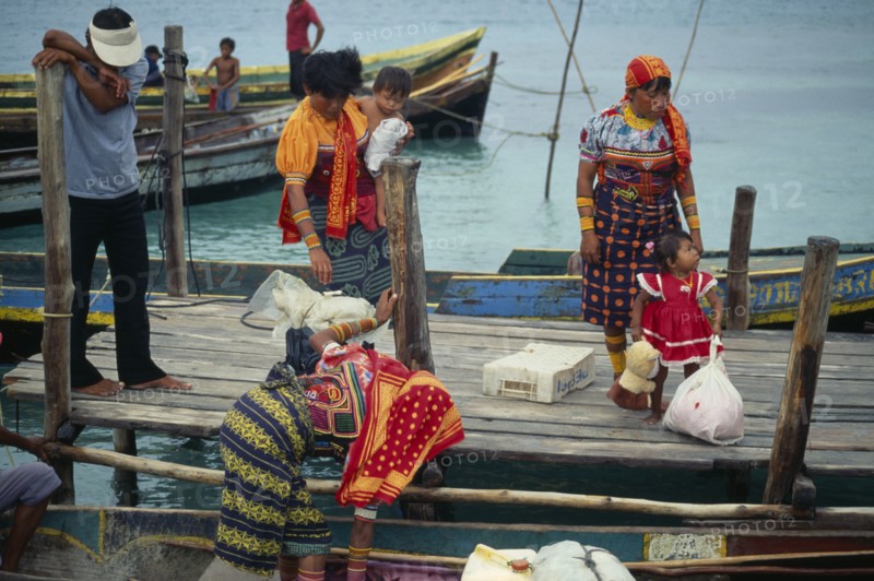 PANAMA, San Blas Islands Tikantiki , Kuna Indians, Kuna Indian women wearing brightly coloured traditional mola panel blouses or dulemolas arriving by canoe at island jetty with children. Cuna Caribbean Niadup American Central America Colored Female Woman Girl Lady Hispanic Kids Latin America Latino Panamanian West Indies