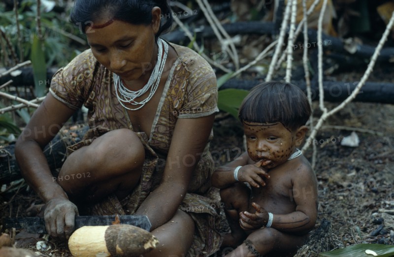 COLOMBIA, North West Amazon, Tukano Indigenous People, Barasana woman peeling manioc roots with small child sitting on ground beside her with painted face and wearing white glass bead necklace and bracelets. Tukano sedentary Indian tribe North Western Amazonia American Children Colombian Columbia Female Women Girl Lady Hispanic Indegent Kids Latin America Latino South America Tukano Female Woman Girl Lady