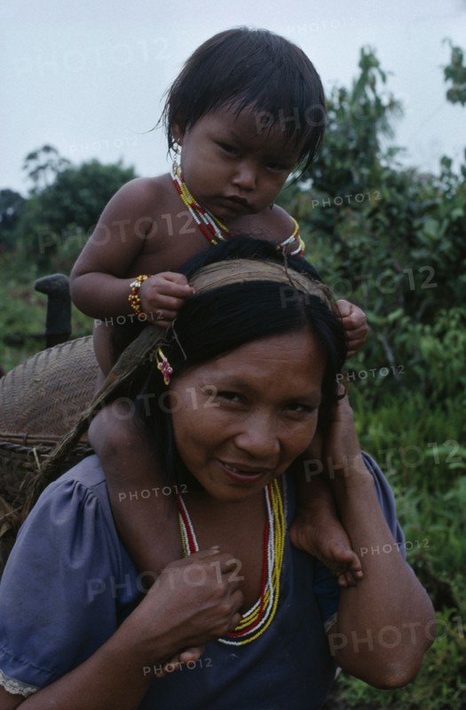 COLOMBIA, North West Amazon, Tukano Indigenous People, Makuna mother carrying her baby son on her shoulders and basket on her back supported by head strap.  Both  wearing necklaces of white red and yellow glass beads. Tukano  Makuna Indian North Western Amazonia family American Babies Colombian Columbia Hispanic Indegent Kids Latin America Latino Mum South America Tukano