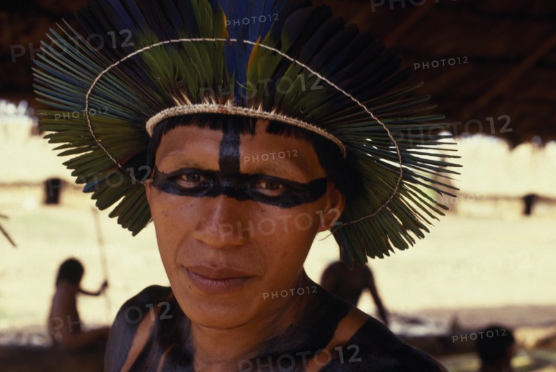 BRAZIL, Mato Grosso, Indigenous Park of the Xingu, Head and shoulders portrait of young male Panara leader wearing ceremonial crown made from green/blue parrot feathers and black face and body paint. Formally known as Kreen-Akrore  Krenhakarore  Krenakore  Krenakarore  Amazon American Brasil Brazilian Indegent Kreen Akore Latin America Latino South America  Formally known as Kreen-Akrore  Krenhakarore  Krenakore  Krenakarore  Amazon American Brasil Brazilian Indegent Kreen Akore Latin America Latino South America Immature One individual Solo Lone Solitary 1 Single unitary Young Unripe Unripened Green