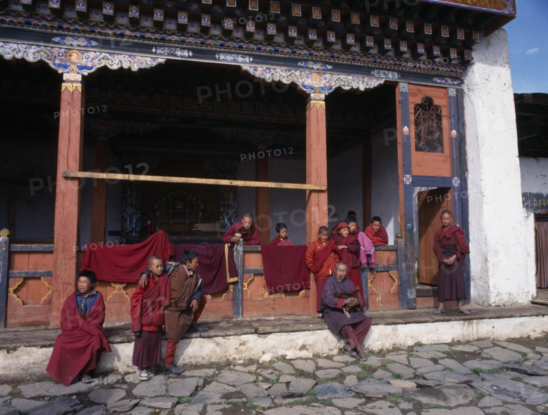 BHUTAN, Gantey Gompa, Group of young novice monks with older man holding cockerel gathered on step outside traditionally painted gompa or monastery.