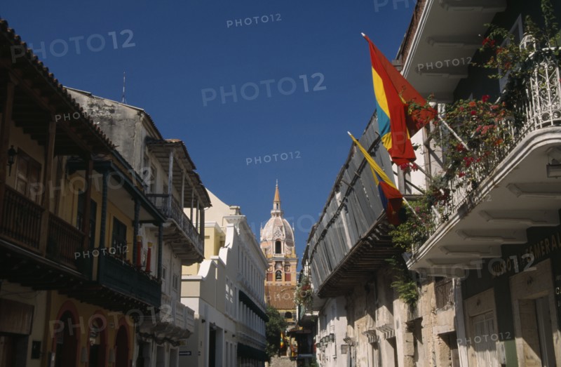 COLOMBIA, Bolivar, Cartagena, "View along Calle San Pedro Claver towards cathedral bell tower and spire.  Narrow street lined with buildings with upper balconies, flags flying in foreground."