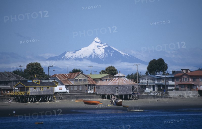 CHILE, Los Lagos, Architecture, Coastal village buildings near Chiloe from sea with snow capped volcanic peak wreathed in cloud behind.