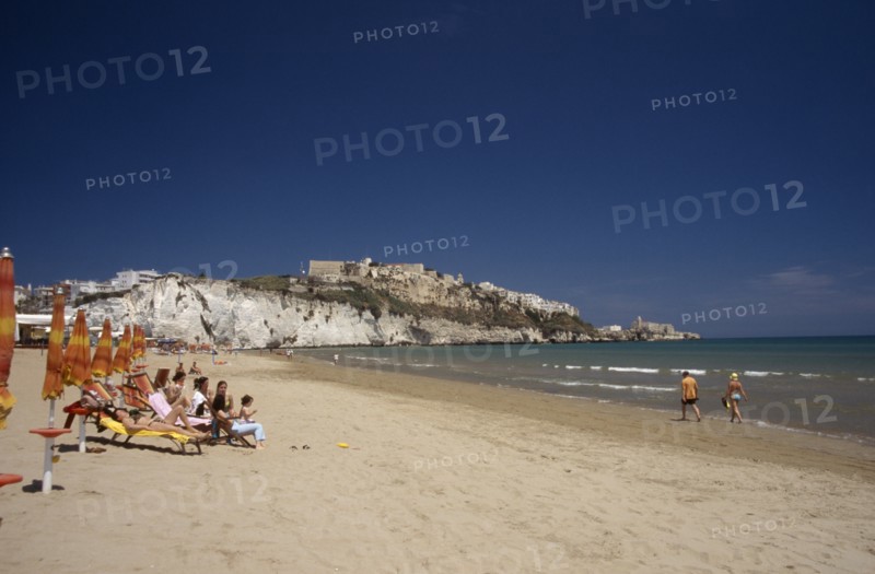 ITALY, Puglia, Foggia, Vieste.  Line of sunbathers on stretch of sandy beach overlooked by castle and apartments on white cliffs beyond.