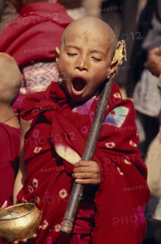 NEPAL, Kathmandu Valley, Bhaktapur, Young monk yawning during a ceremony and wearing red robes outside a small monastery in the back streets