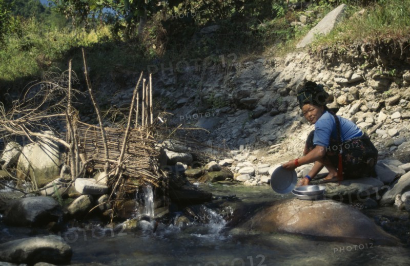 NEPAL, Manaslu Circuit Trek, Arughat, Woman washing pots beside a fish trap in a tributary river near Arughat Village