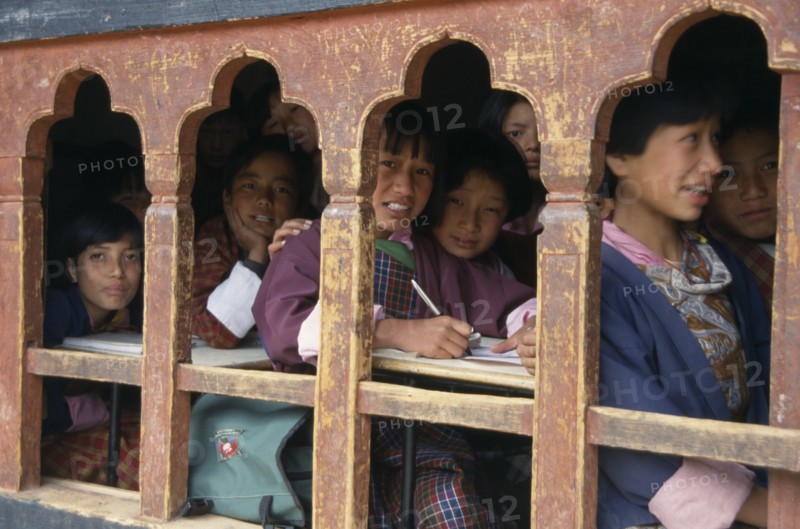 BHUTAN, Thimphu, "School children in class writing at desks framed in line of low set, arched, glassless windows."