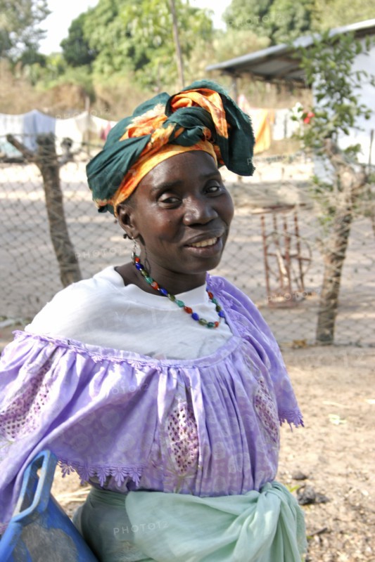 GAMBIA, Western Gambia, Tanji, Tanji village.  Three-quarter portrait of woman wearing traditional head tie or scarf and bead necklace posing in front of her home compound.