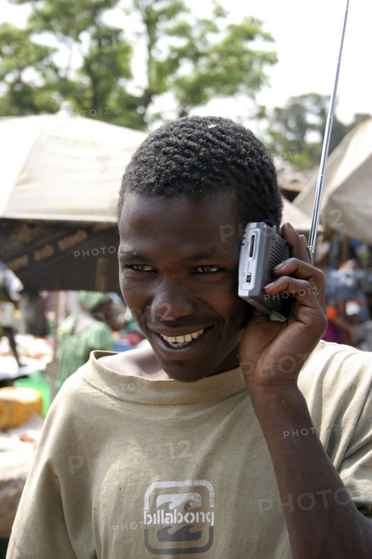 GAMBIA, Atlantic Coast, Banjul, "Albert Market, Russell Street.  Young African man smiling  while listening to a radio which he is holding next to his ear."