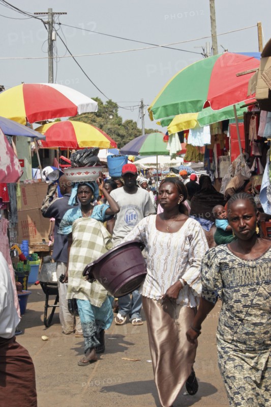 GAMBIA, Western Gambia, Serekunda, "Bakau Market, Atlantic Road.  Busy market scene with crowds of people, and roadside stalls beneath colourful striped sun umbrellas. "