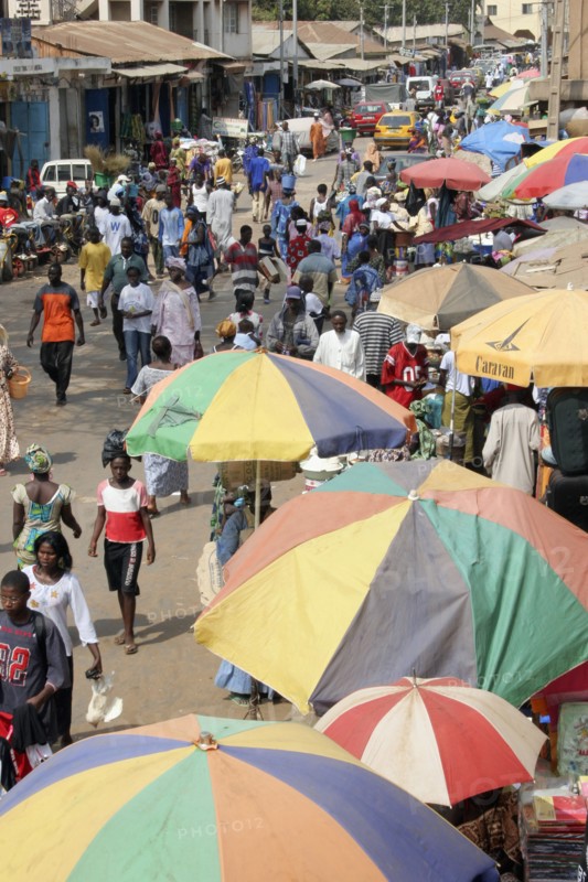 GAMBIA, Western Gambia, Serekunda, "Bakau Market, Atlantic Road.  Busy city street lined with stalls beneath colourful striped sun umbrellas.  Crowds of people, traffic and shop fronts open to the street.                   "