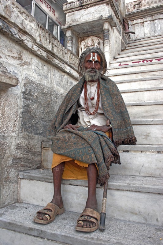 INDIA, Rajasthan, Udaipur, Elderly male Hindu beggar sitting on steps outside the Jagdish Temple wrapped in blanket shawl with painted forehead and grey beard.