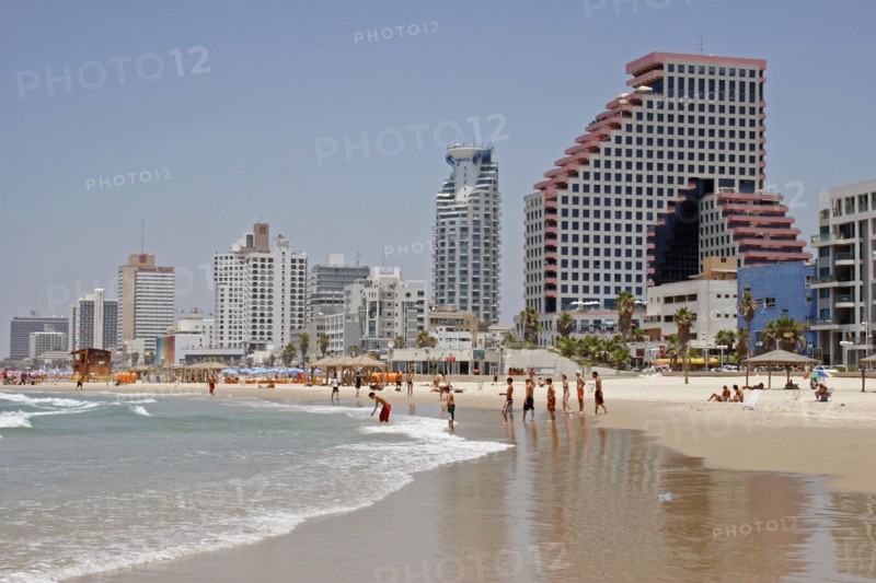 ISRAEL, Tel Aviv, "Beach with people sunbathing and at waters edge overlooked by high rise, modern city buildings reflected on wet sand."