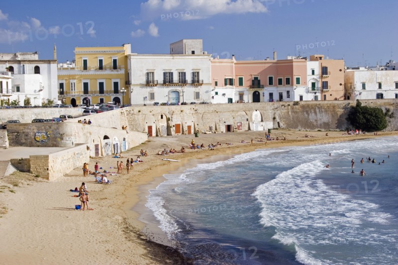ITALY, Puglia, Gallipoli, View of old city with traditional houses beside sea wall overlooking curving sandy beach with people sunbathing and swimming