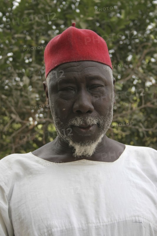 GAMBIA, Western Gambia, Tanji, Tanji Village.  Head and shoulders portrait of Muslim man with white trimmed beard wearing white shirt and red cap.