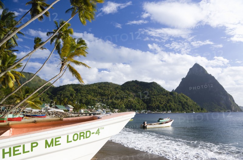WEST INDIES, St Lucia, Soufriere, Fishing boats on the beach lined with coconut palm trees with the town and the volcanic plug mountain of Petit Piton beyond. A fishing boat in the foreground with the words Help Me Lord written on the bow