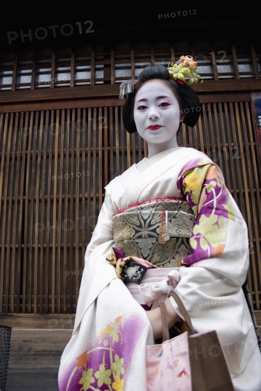 JAPAN, Honshu, Kyoto, "Gion District, the neighbourhood where Geisha live and perform.  Three-quarter portrait of smiling Maiko, or apprentice Geisha with hair pinned up with decorative ornaments, white facial make up, red painted lips and wearing traditional kimono of pale, flower patterned silk. "