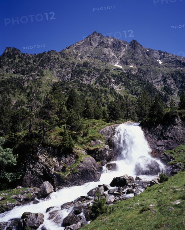 FRANCE, Midi-Pyrenees, Hautes-Pyrenees, Vallee du Lutour.  Jagged peaks of Soum det Guingays 2451 m / 8027 ft above white waterfall.  Outcrops of rock scattered over hillside amongst trees.