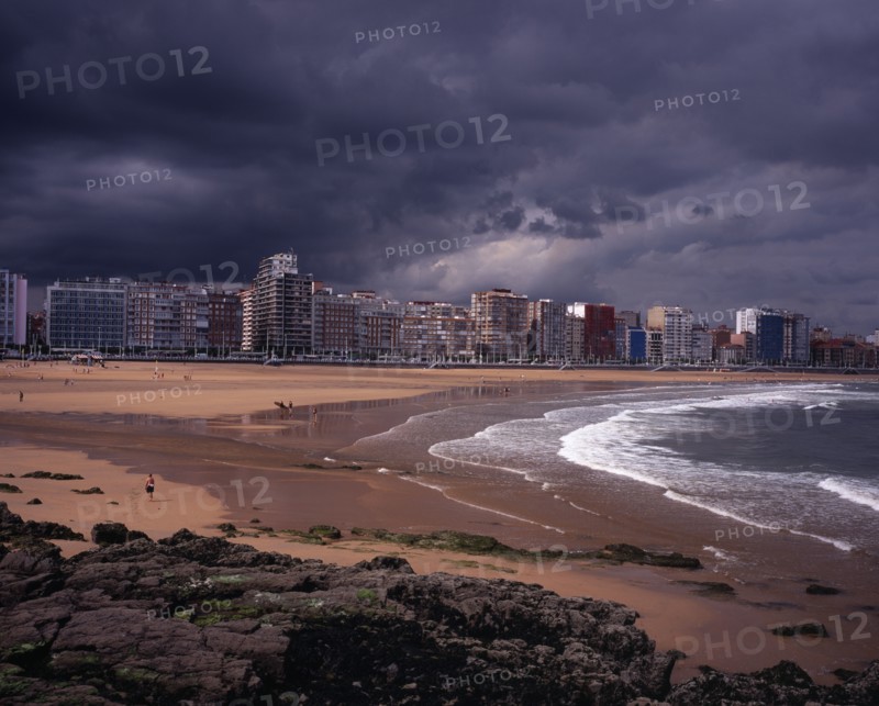 SPAIN, Asturias, Gijon, "High rise city buildings overlooking beach with people in water, on beach and surfing.  Grey, cloudy sky with approaching storm.  Rocks and seaweed in foreground."
