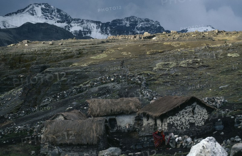 BOLIVIA, Northern Andes, Antaquilla , Collpa Huata Llama herders settlement of stone houses with thatched roofs. A woman wearing traditional red textiles standing next to stone wall. Snow covered mountains behind. Near Peru