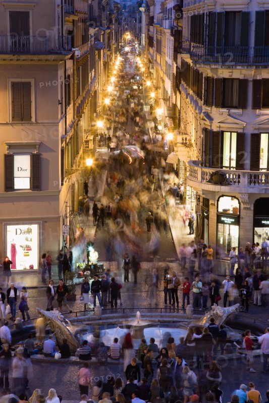 ITALY, Lazio, Rome, The Via dei Condotti the main shopping street busy with people illuminated at night seen from the Spanish Steps with seated tourists and the Fontana della Barcaccia in the foreground