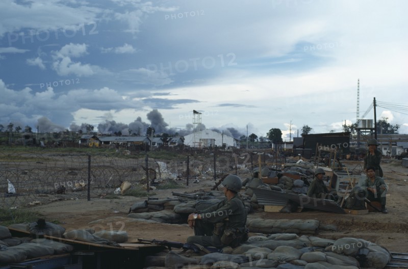 VIETNAM, Central Highlands, Kontum, Vietnam War. Siege of Kontum. American B-52 bombing behind Kontum. Montagnard soldiers sitting in camp within a barbed wire perimeter watching plumes of grey smoke rising into the sky