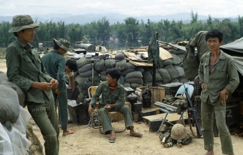 VIETNAM, Central Highlands, Kontum, "Vietnam War. Siege of Kontum. Montagnard soldiers gathered in base within a barbed wire perimeter. Young soldier sitting on a chair holding a medical syringe. Walls of sand bags, makeshift shelters, tools and artillery."