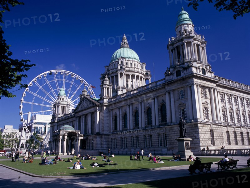 IRELAND, North, Belfast, People sitting or lying in the sun in the grounds of City Hall with The Wheel of Belfast part seen behind.