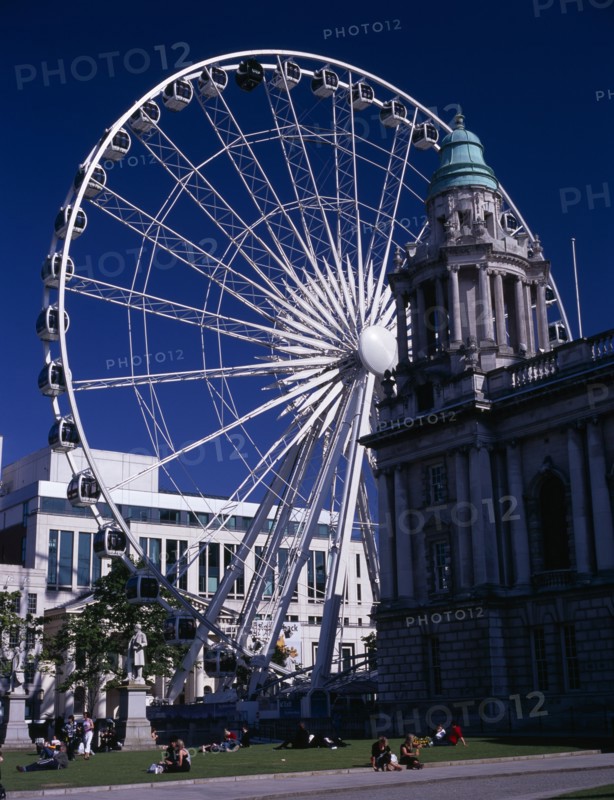 IRELAND, North, Belfast, People sitting or lying in the sun in the grounds of City Hall with The Wheel of Belfast part seen behind.