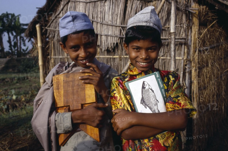 BANGLADESH, Shariatpur, "Three-quarter portrait of two, smiling young boys carrying books and writing board standing outside thatched hut."