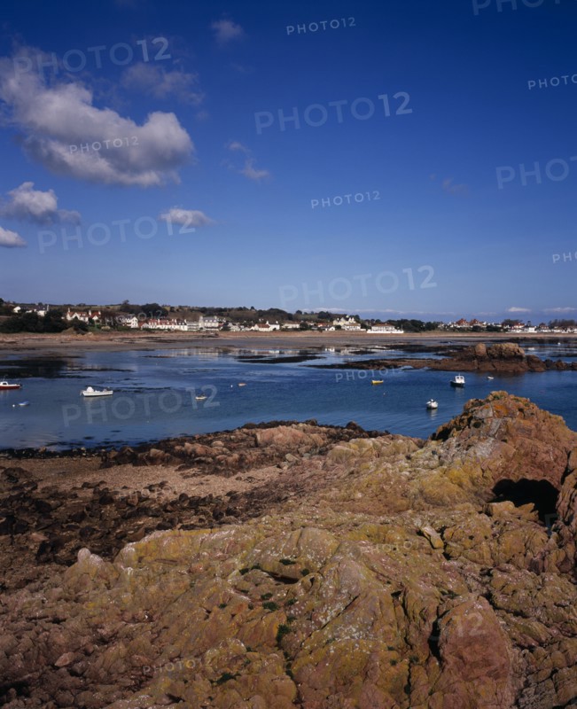 UNITED KINGDOM, Channel Islands, Jersey, Grouville. View across rocky foreshore with small boats on the sea towards Rocque Village