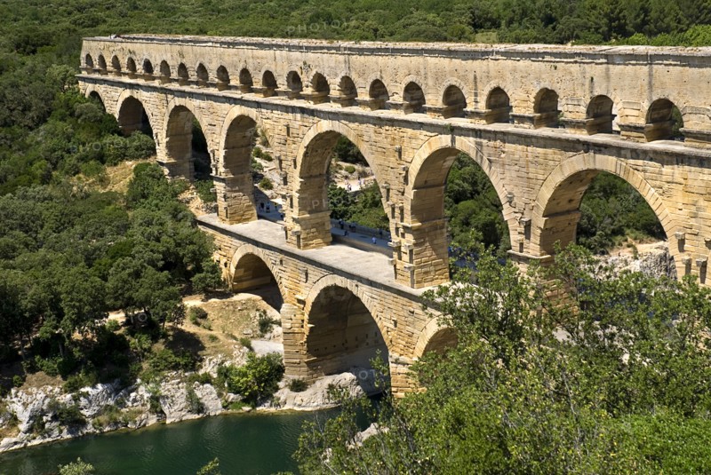 FRANCE, Provence Cote d’Azur, Gard, Pont du Gard Roman aqueduct from a high vantage point on the western side showing three tiers of continuous arches spanning river and visitors on bridge.