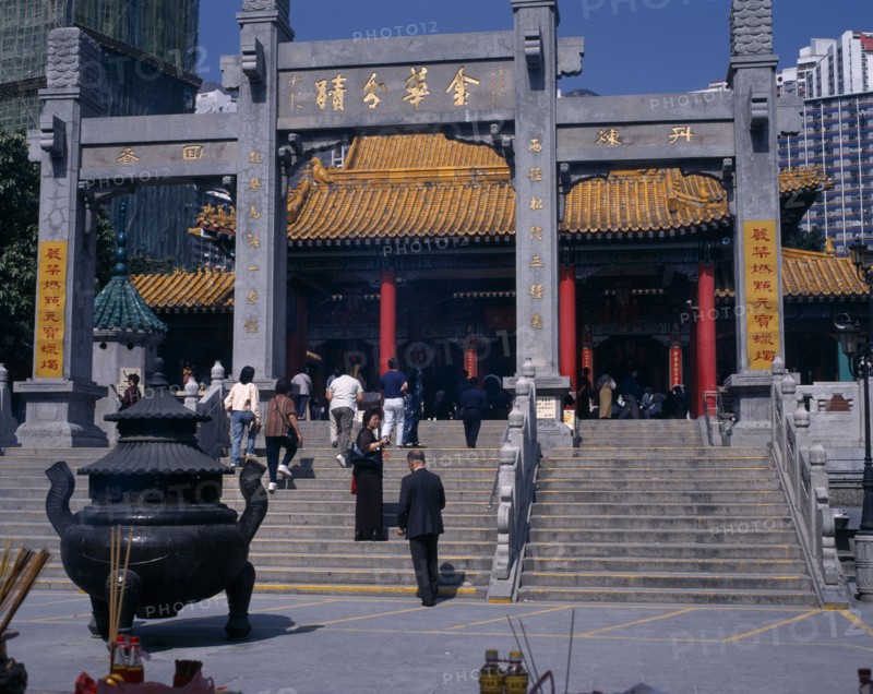 CHINA, Hong Kong, Kowloon, "Wong Tai Sin taoist temple established in 1921.  Visitors on flight of steps to entrance built in traditional Chinese style with red interior pillars and pagoda style roof.   Incense, offerings and burner in foreground. "