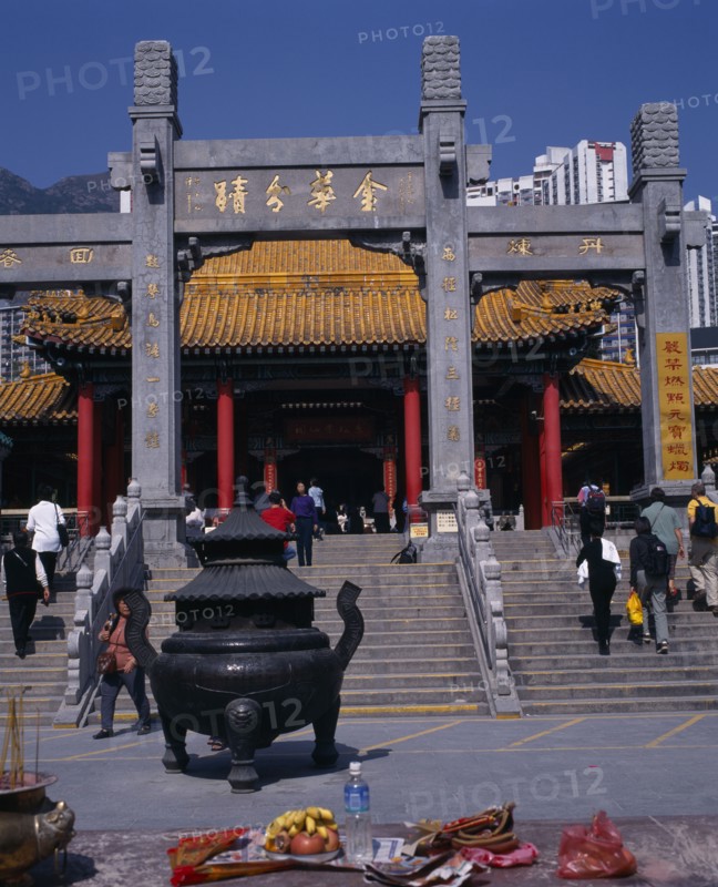 CHINA, Hong Kong, Kowloon, "Wong Tai Sin taoist temple established in 1921.  Visitors on flight of steps to entrance built in traditional Chinese style with red interior pillars and pagoda style roof.   Incense, burner and offerings in foreground. "