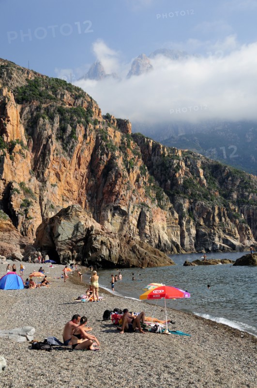 FRANCE, Corsica, Golfe Di Porto, "Plage De Bussaglia, sandy beach with sunbathers & umbrellas. Rocky cliffs & cloud on hills"