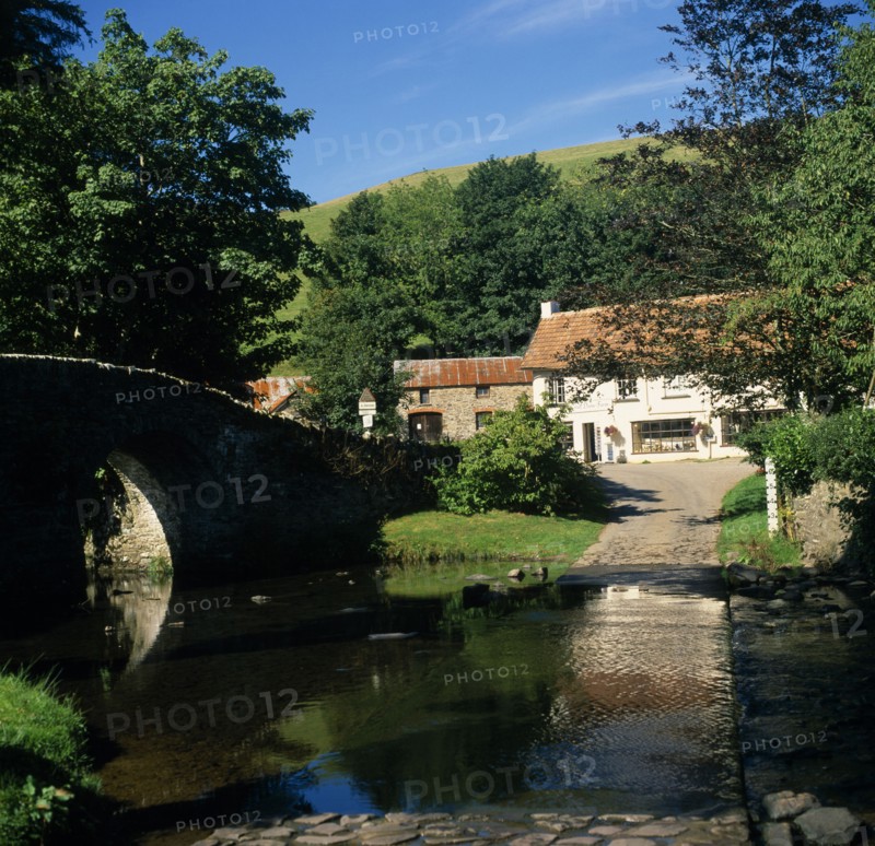 ENGLAND, Devon, Exmoor, Malmsmead on Badgworthy Water. Arched stone bridge and ford across river leading to ‘Lorna Doone Farm’ shop White painted building with tiled roof  and outbuildings.