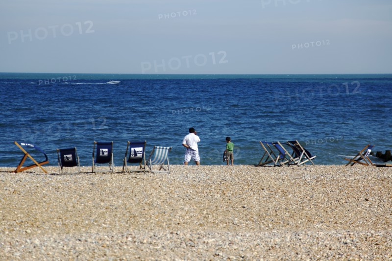 ENGLAND, East Sussex, Eastbourne, Man and young boy on the beach with deck chairs blowing in the wind. Speed passing by in the distance.