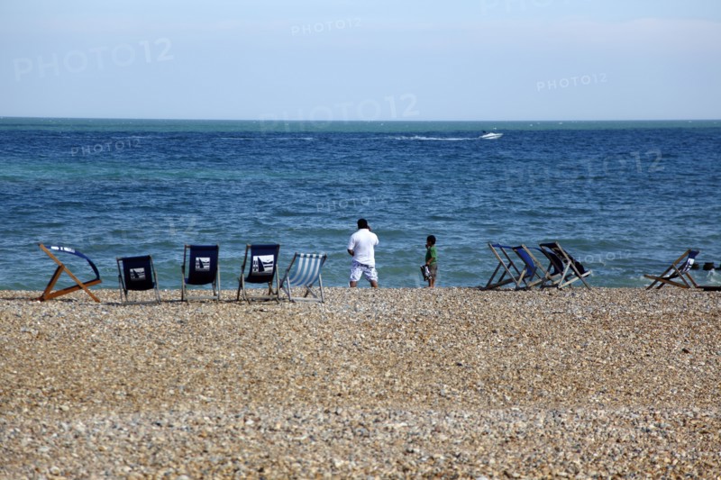 ENGLAND, East Sussex, Eastbourne, Man and young boy on the beach with deck chairs blowing in the wind. Speed passing by in the distance.