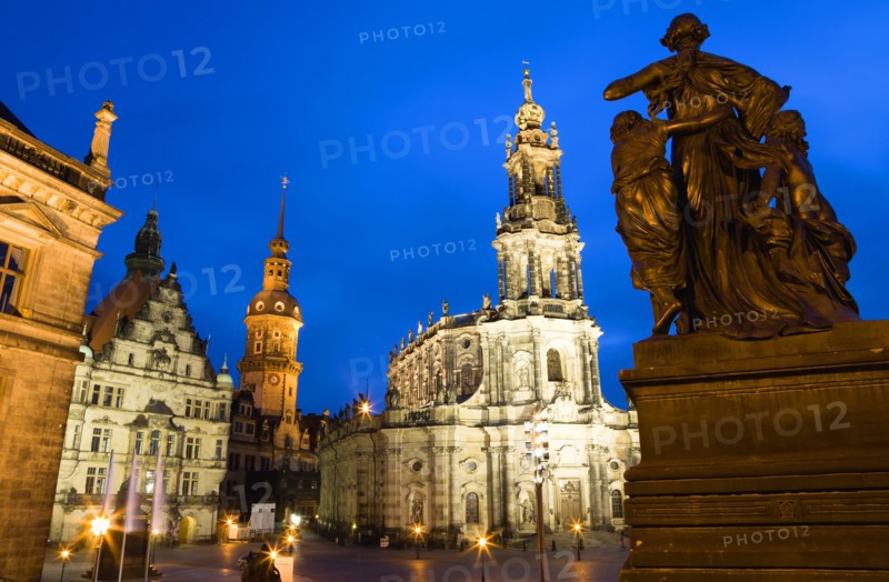 GERMANY, Saxony, Dresden, The 18th Century Hofkirche Catholic Cathedral of Saint Trinitatis and the illuminated tower of the Residenzschloss and the Grunes Gewelbe or Green Vault a museum that is part of Dresden Castle with a statue of a woman and children in the foreground at sunset.