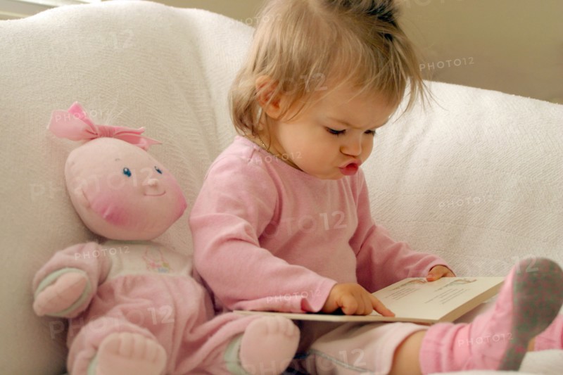 EDUCATION, Children, Reading, Infant girl dressed in pink reading book whilst mouthing the words and following text on the page with her finger.  Pink doll propped up on chair beside her.