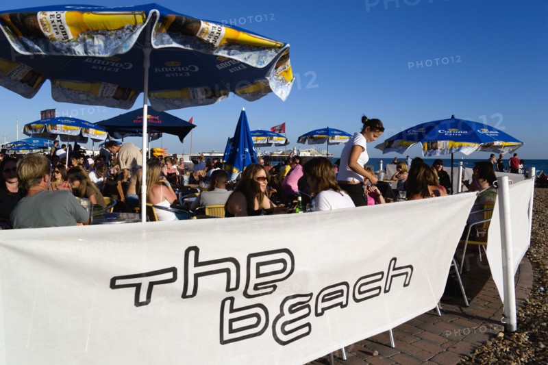 ENGLAND, East Sussex, Brighton, People sitting under sun shade umbrellas at tables on the promenade outside The Beach Bar with The Brighton Pier beyond.