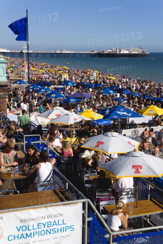 ENGLAND, East Sussex, Brighton, People sitting under sun shade umbrellas at tables on the promenade outside bars and restaurants with Brighton Pier and people on the shingle pebble beach beyond.