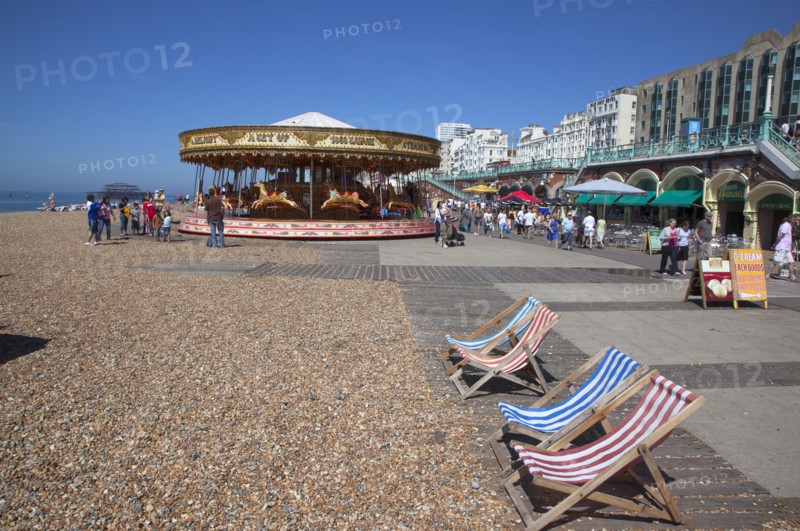 England, East Sussex, Brighton, Deck chairs and carousel on the pebble beach promenade.