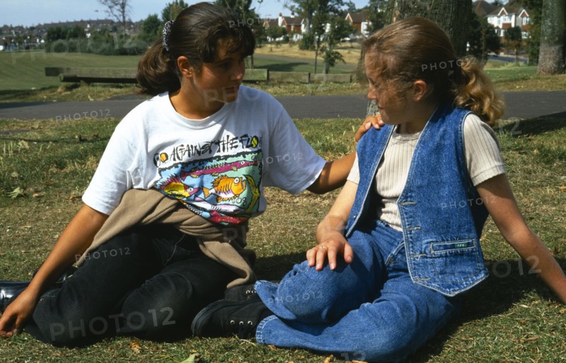 England, East Sussex, Brighton, One teenage girl reassuring another young girl sitting on the grass in Hove Park.